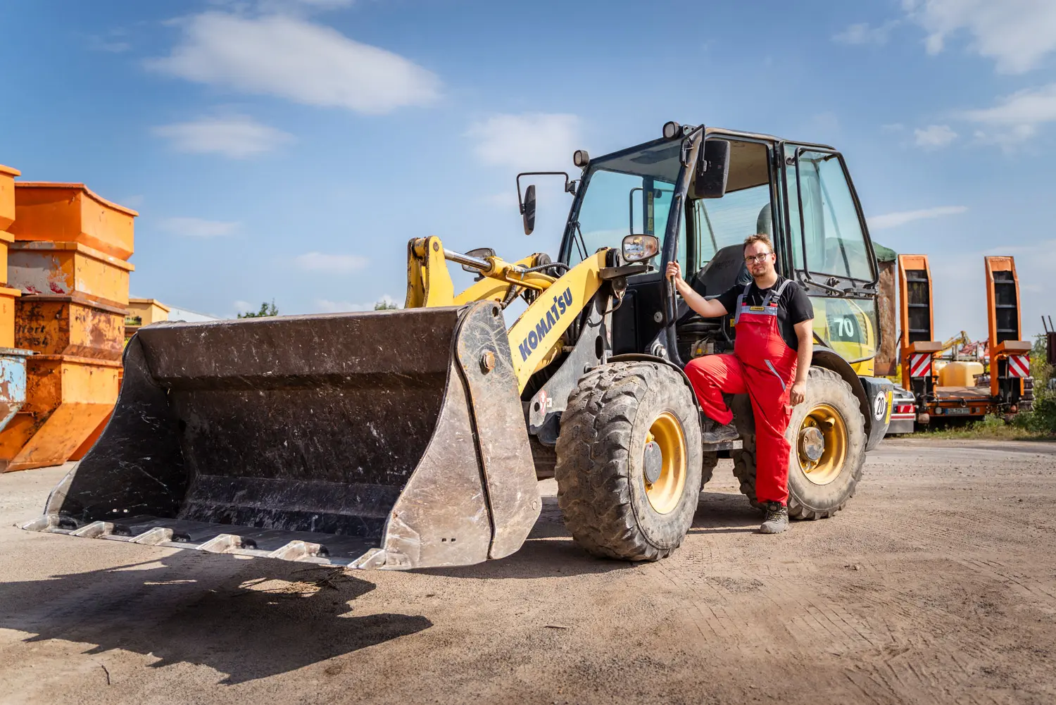 Mitarbeiter in roter Latzhose steht neben einem gelben Radlader auf einem Baustellengelände.
