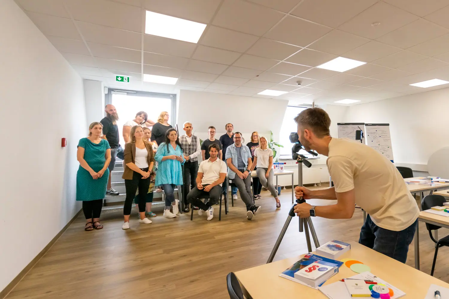 Fotograf richtet in einem Seminarraum eine Kamera auf eine Gruppe von Mitarbeitenden, die für ein Teamfoto posieren.