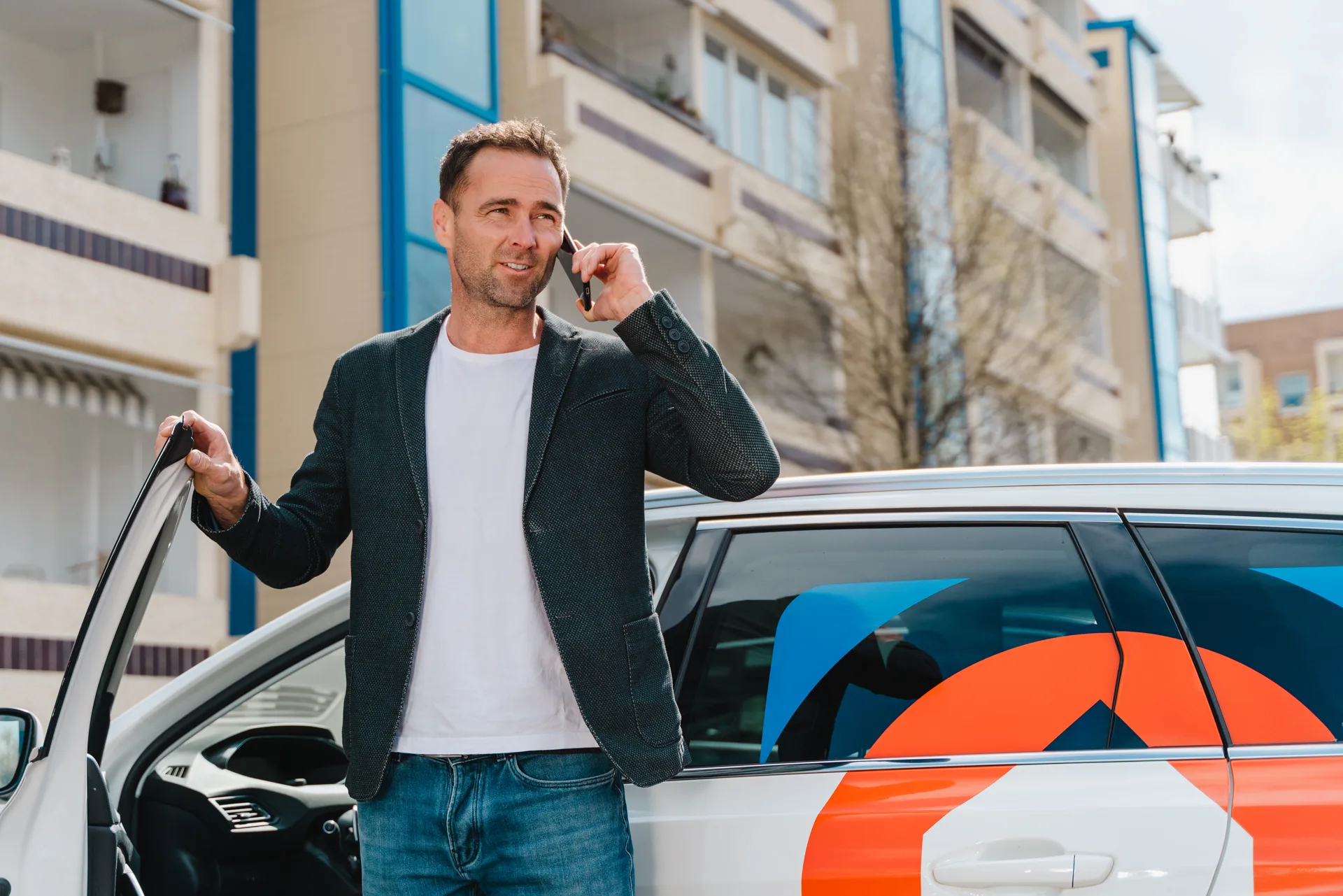 Mann steht neben einem Auto mit Firmenlogo, hält ein Handy ans Ohr und spricht, im Hintergrund Wohngebäude.