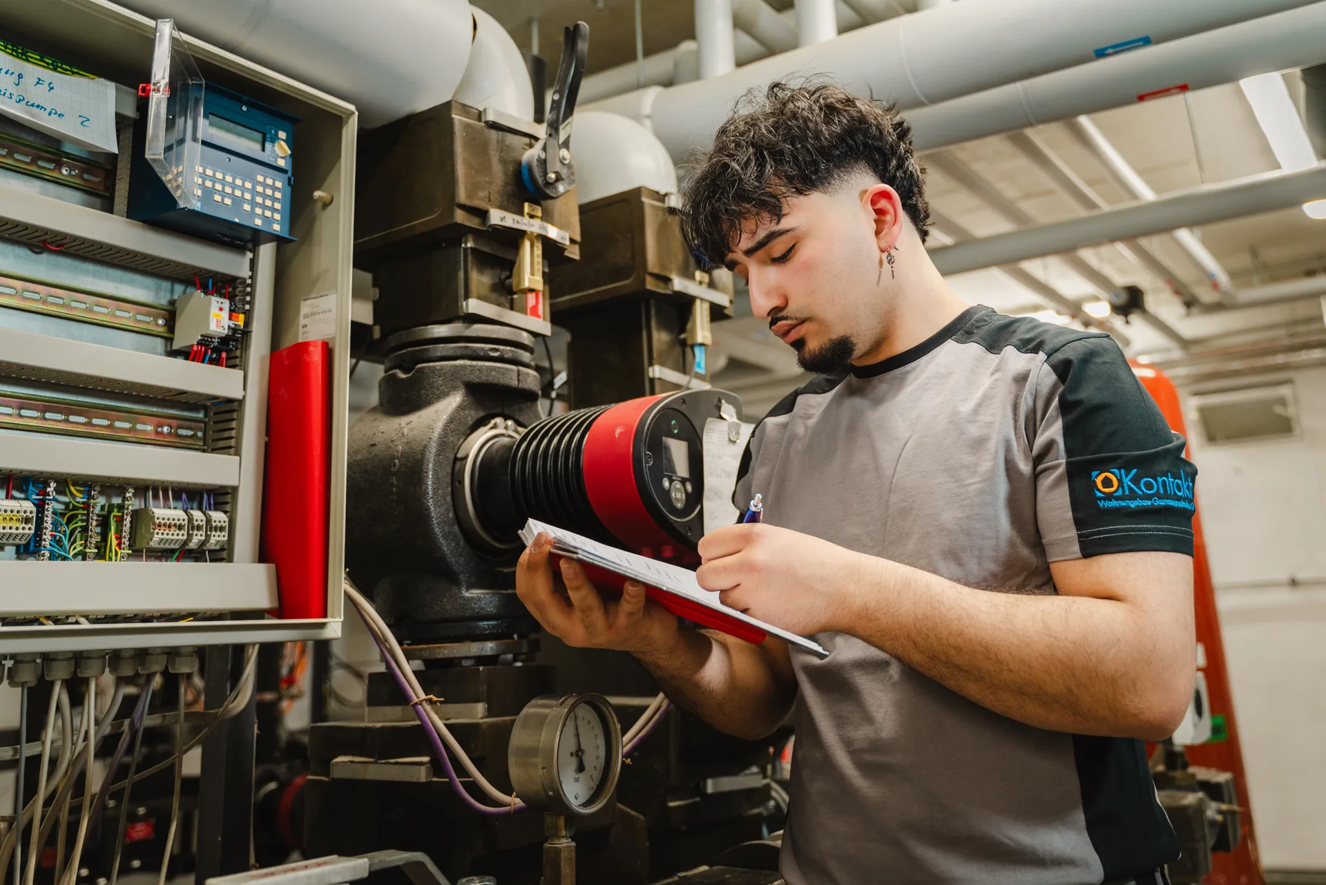 Techniker überprüft Heizungsanlage und notiert Messwerte. Im Hintergrund Rohrleitungen und Steuerungskästen.
