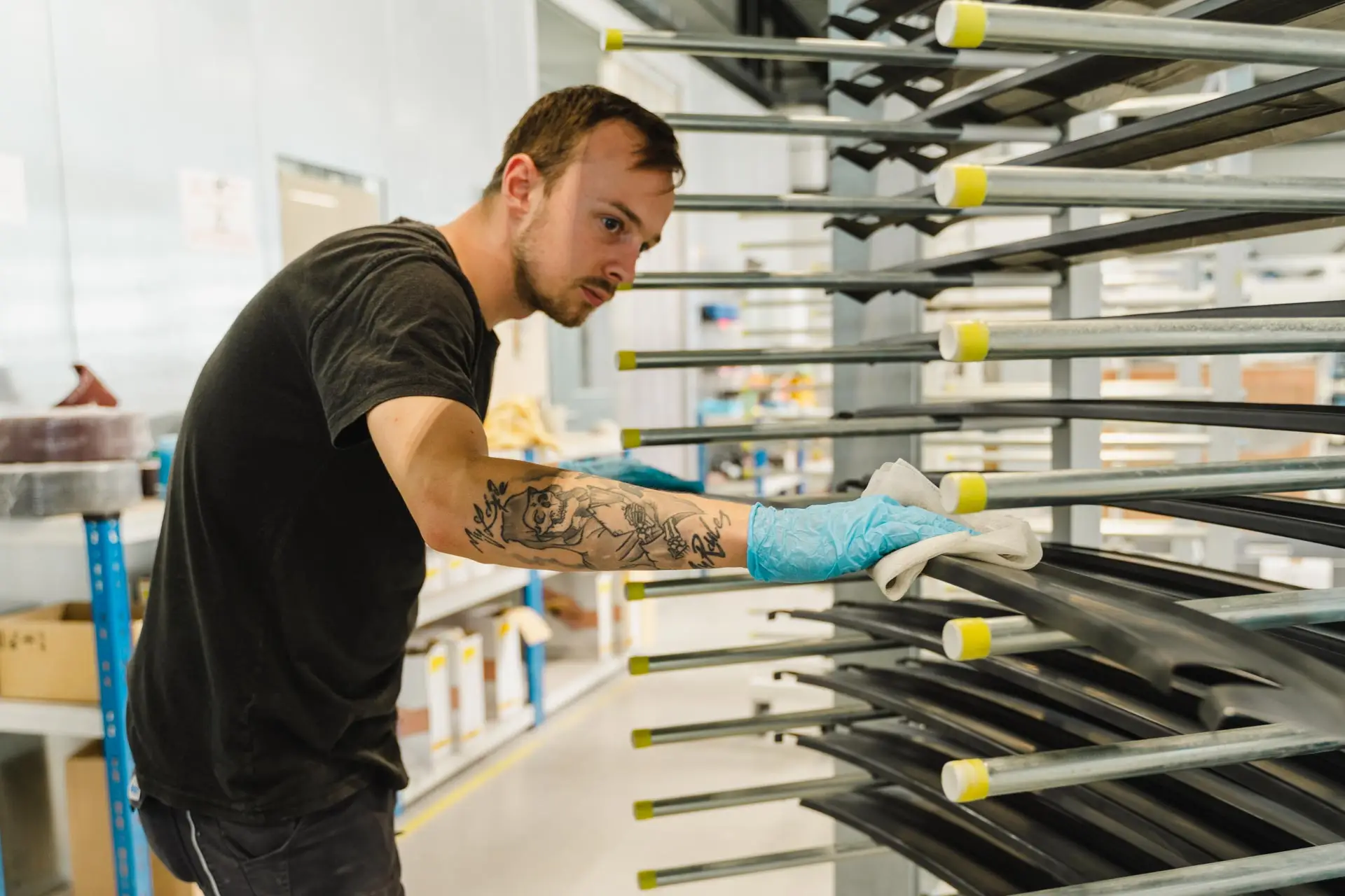 Mann mit tätowiertem Arm reinigt sorgfältig schwarze Werkstücke auf einem Metallregal in einer Werkhalle.