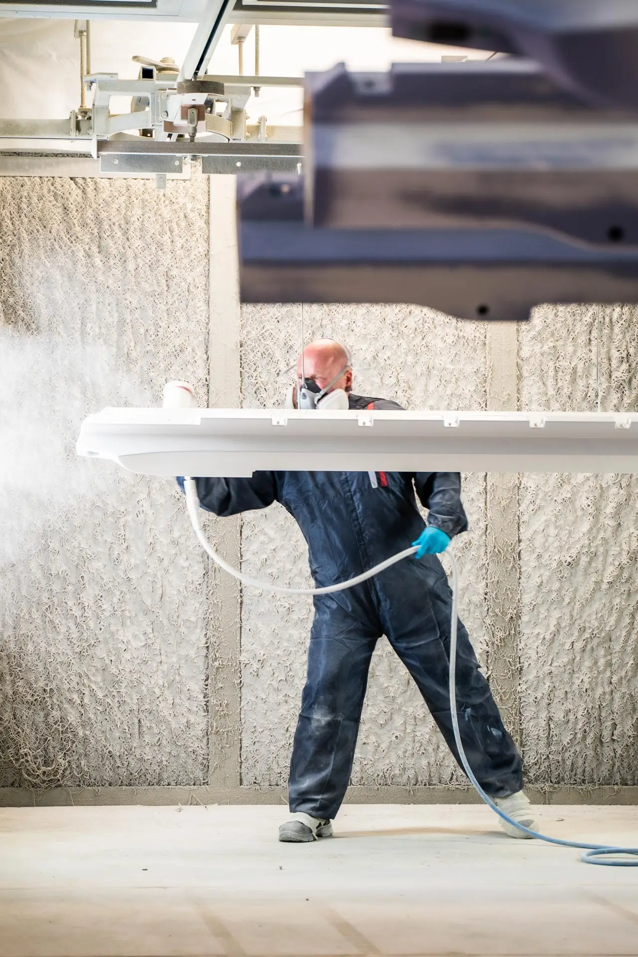 Mitarbeiter trägt mit einer Lackierpistole weiße Farbe auf ein großes Werkstück in einer Spritzkabine auf.