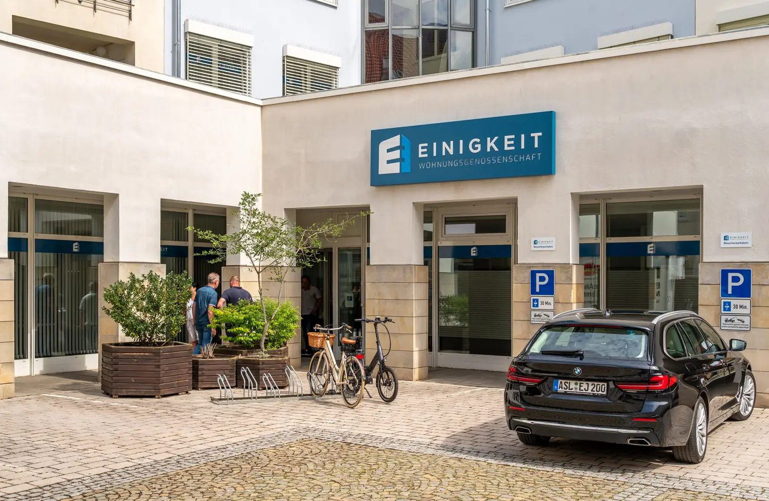 Außenansicht der Geschäftsstelle der Einigkeit Wohnungsgenossenschaft mit Logo, Fahrrädern und parkendem Auto im Innenhof.
